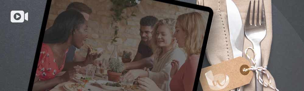 A tablet displays a group of five people enjoying a meal together, smiling and engaged in conversation, set in a warm, well-lit dining environment with stone walls.
