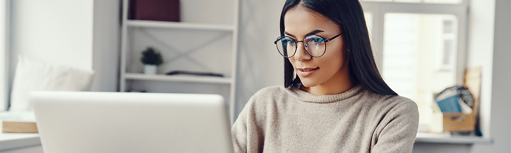 A young woman wearing glasses is focused on her laptop while sitting in a cozy, well-lit workspace. Shelves and soft furnishings create a comfortable atmosphere.