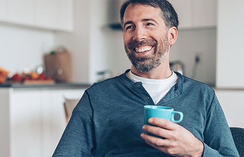 A man with a beard is smiling while holding a blue mug. He is seated in a cozy kitchen with light-colored cabinetry and some items in the background.