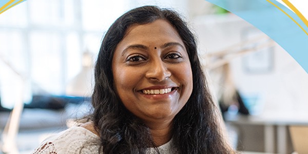 A woman with long, dark hair smiles warmly while seated in a brightly lit indoor space. She wears a white top and is surrounded by a modern, open office environment.