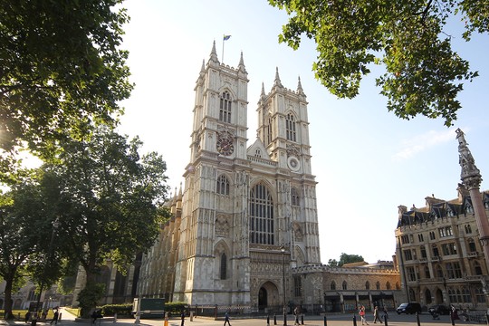 Tall Gothic cathedral featuring ornate towers and a large entrance, surrounded by trees and people walking on a street in bright daylight, with a flag atop one tower.