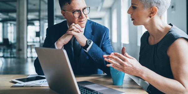 A man in a suit and glasses listens attentively as a woman in a sleeveless dress gestures while speaking. They sit at a table with a laptop and office materials in a modern office setting.