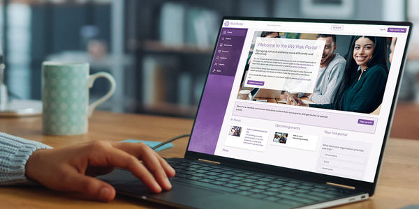 A laptop rests on a desk. A hand interacts with the keyboard while the screen displays a web portal titled "BW Risk Portal," featuring resource links and event details. A patterned mug sits nearby.