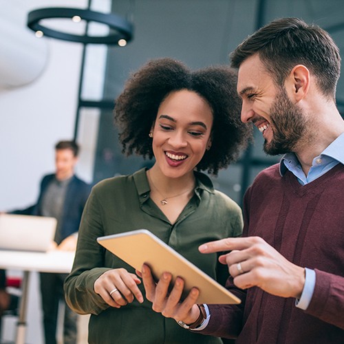 A tablet is held between two smiling colleagues, who are engaged in discussion. In the background, two others work at a desk, indicating a collaborative office environment.