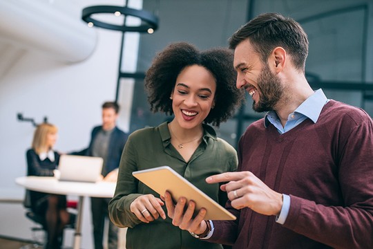 A tablet is held between two smiling colleagues, who are engaged in discussion. In the background, two others work at a desk, indicating a collaborative office environment.