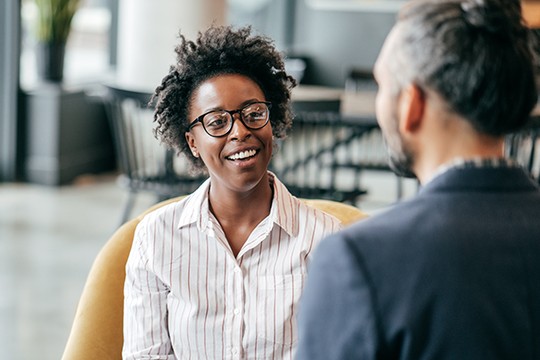 A woman with curly hair and glasses smiles while engaging in conversation with a man. They are seated in a modern, cozy environment with stylish furniture in the background.