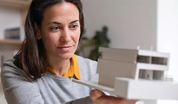 A woman examines a detailed architectural model with focus, holding it carefully in her hands. The background is a softly lit room with minimal decoration, emphasizing her concentration.