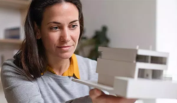 A woman examines a detailed architectural model with focus, holding it carefully in her hands. The background is a softly lit room with minimal decoration, emphasizing her concentration.