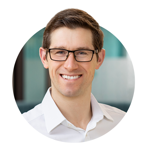 A young man with short hair and glasses smiles confidently. He is wearing a white button-up shirt. The background features blurred office elements, suggesting a professional environment.