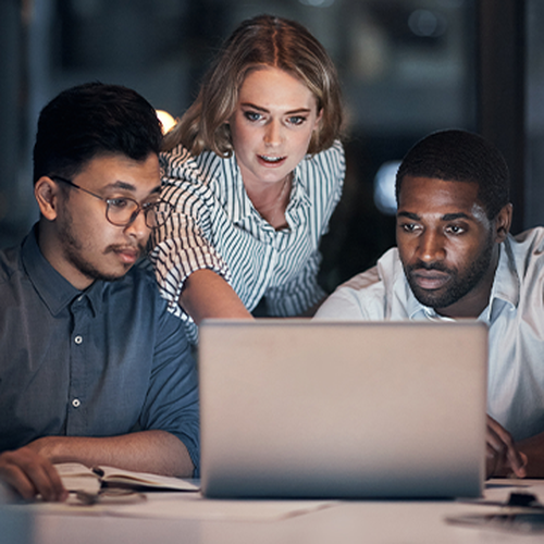 Three people focus on a laptop screen in a dimly lit office. One woman stands, pointing at the screen, while two men sit, looking engaged and thoughtful. The atmosphere is collaborative.