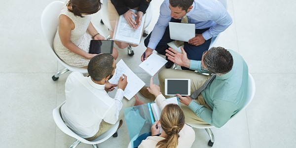 A group of six people sit in a circle, collaborating on documents and devices, in a well-lit modern space with a light-colored tiled floor.