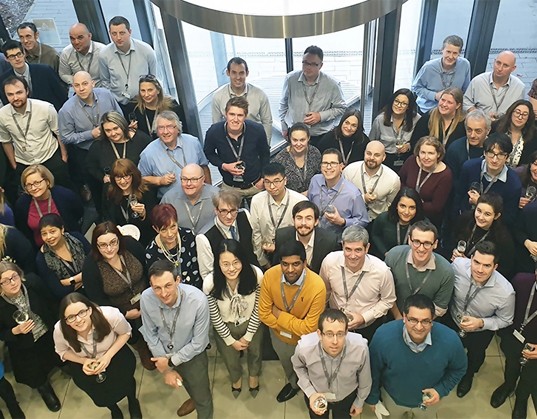 A large group of people stands together in a well-lit, modern space with large windows, some holding drinks and smiling, creating a celebratory atmosphere.