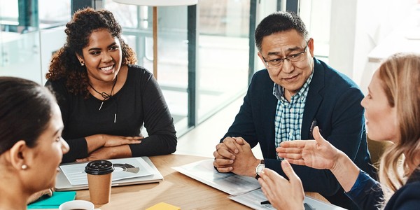 A group of five professionals engages in discussion around a large table. One woman gestures while speaking, and others listen attentively in a bright, modern office setting.