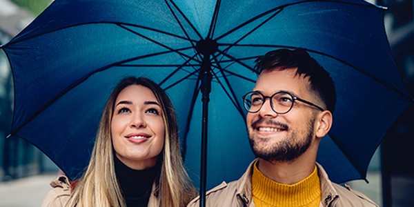 Two people stand under a large blue umbrella, smiling and looking up. They are dressed in light jackets, and the environment suggests an urban setting with smooth surfaces and soft lighting.