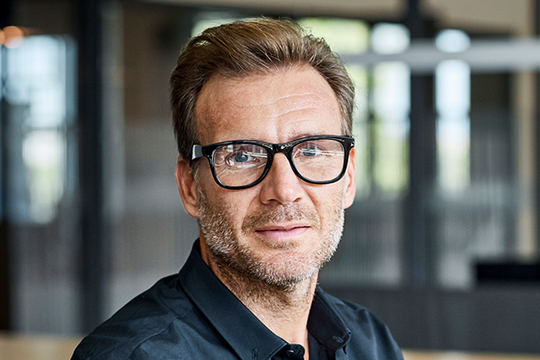 A man with short, tousled hair is wearing glasses and a black shirt, looking directly at the camera with a neutral expression. The background features blurred office elements.