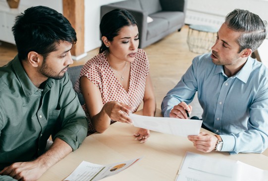 Three adults sit around a table, discussing a document. One person points at the paper while the others listen attentively. The setting is a modern, well-lit office space.