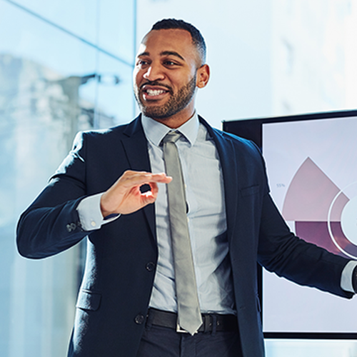 A well-dressed man gestures while presenting in front of a screen displaying a pie chart, surrounded by large windows that let in natural light, suggesting a business environment.