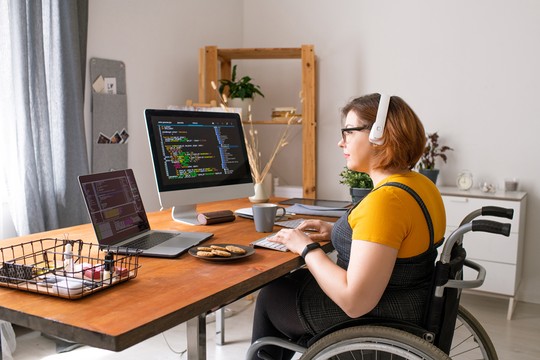 A woman in a wheelchair types on a laptop and desktop, wearing headphones. Cookies and a coffee cup sit on a wooden table in a bright, modern home office with plants.