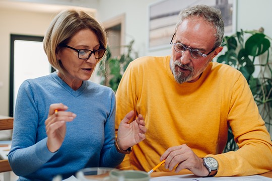 A woman in a blue sweater and glasses gestures while discussing with a man in a yellow sweater. They are seated at a table with documents and plants in a bright room.