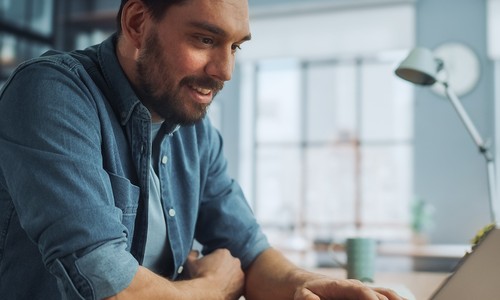 A man uses a laptop, smiling as he engages with it. He sits in a bright, modern workspace with large windows, greenery, and wooden shelves in the background.