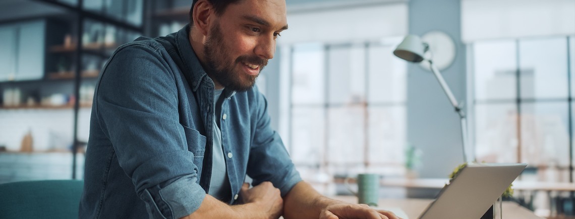 A man uses a laptop, smiling as he engages with it. He sits in a bright, modern workspace with large windows, greenery, and wooden shelves in the background.