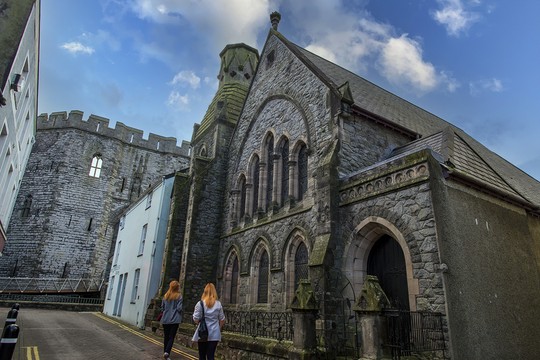 Two individuals walk along a narrow street lined with historic stone buildings, passing a large church with decorative arches and a nearby castle-like structure against a partly cloudy sky.
