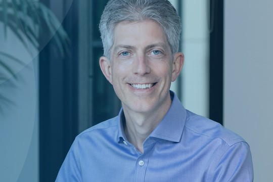 A man with gray hair smiles while sitting in an office. He wears a light blue shirt. In the background, there are plants and large windows indicating a professional environment.
