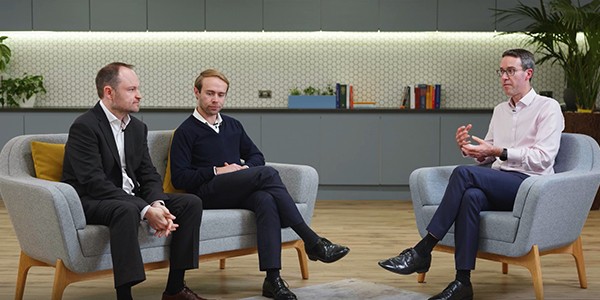 Three men sit on a light gray sofa in a modern, well-lit room with plants and bookshelves. One man gestures while discussing, while the others listen attentively.