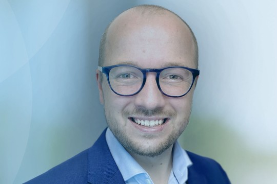 A man with glasses and a suit smiles. He is set against a soft-focus blue background, conveying a professional and friendly atmosphere, suggesting a corporate or formal context.
