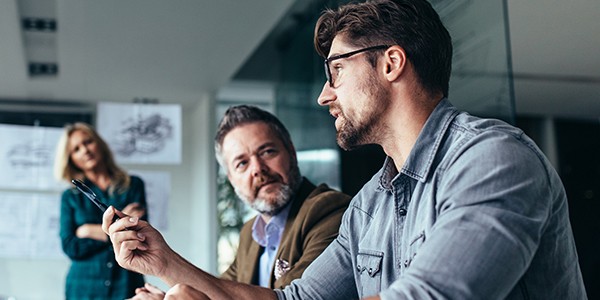 A man holds a pen, engaging in discussion with another man, while a woman stands in the background. They are in a modern meeting room with whiteboard sketches visible.