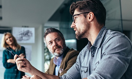 A man holds a pen, engaging in discussion with another man, while a woman stands in the background. They are in a modern meeting room with whiteboard sketches visible.