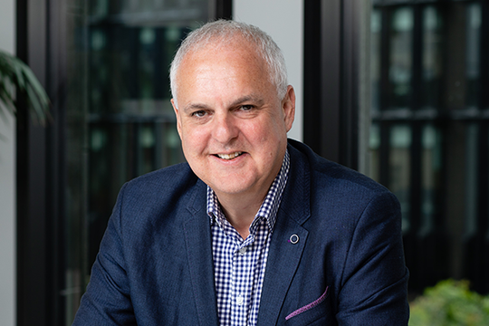 A man with short gray hair and a friendly smile sits confidently at a table. He wears a blue blazer over a checked shirt, surrounded by large windows in a modern office environment.