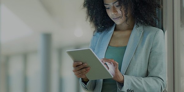 A woman with curly hair is focused on a tablet, standing against a wall in a modern, well-lit hallway. She wears a light gray blazer over a green top.