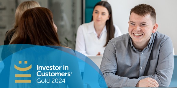 A young man smiles while engaged in conversation with colleagues in a modern office setting. The context includes a bright space with casual seating and a glass wall. The text reads: "Investor in Customers® Gold 2024."