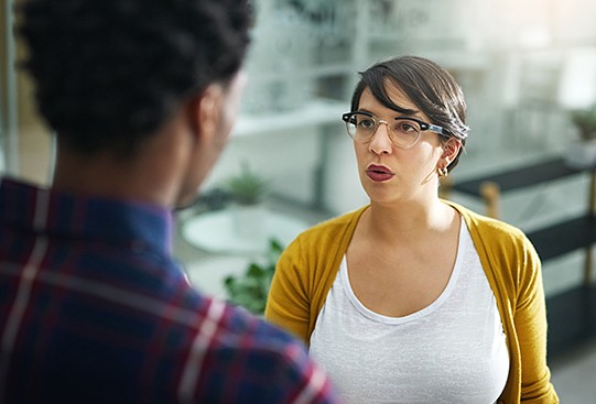 A woman with glasses and a yellow cardigan speaks assertively to a man, engaged in conversation in a bright, modern workspace filled with greenery and soft furnishings.