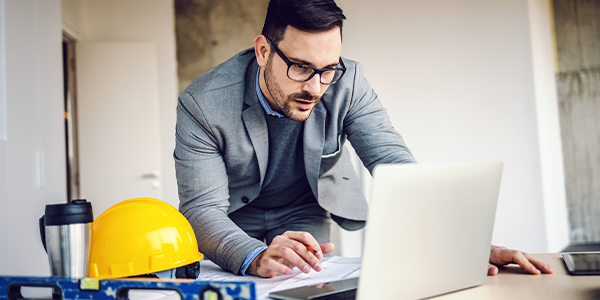 A man in a suit and glasses is leaning over a laptop, examining documents. A yellow hard hat and a level are on the table, indicating a construction or engineering environment.