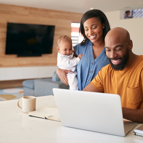 A man smiles at a laptop while a woman holds a baby. They are in a modern living room with wooden accents, a couch, and a coffee cup on the table.