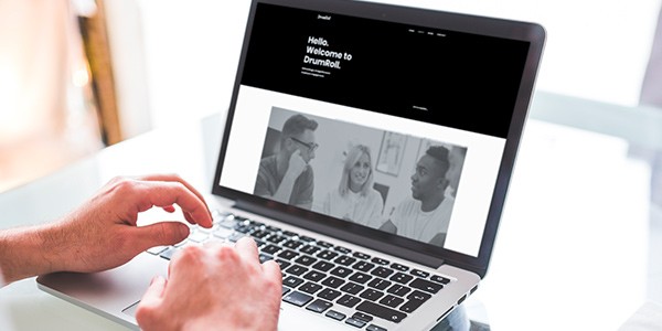 A person types on a laptop displaying a website with a greeting. The context features a bright indoor space, suggesting a casual, friendly environment. Text includes: "Hello. Welcome to DrumRoll."
