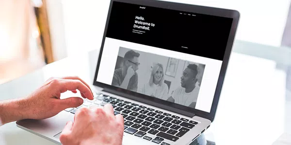 A person types on a laptop displaying a website with a greeting. The context features a bright indoor space, suggesting a casual, friendly environment. Text includes: "Hello. Welcome to DrumRoll."