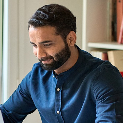 A man with a beard wears a blue shirt while sitting at a desk, smiling as he interacts with a laptop. Bookshelves filled with books are visible in the background.