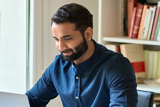 A man with a beard wears a blue shirt while sitting at a desk, smiling as he interacts with a laptop. Bookshelves filled with books are visible in the background.