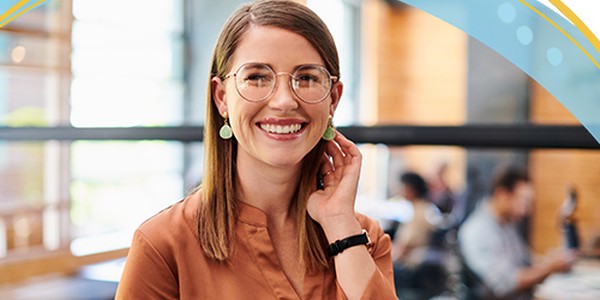 A woman with glasses and earrings smiles while playing with her hair. She stands in a café with a wooden interior, while other people are seated in the background.