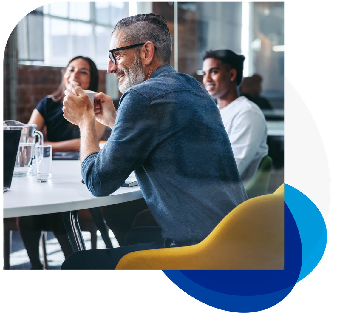 A man with glasses smiles while holding a cup, engaged in conversation with others at a modern table in a well-lit, airy meeting room featuring brick walls and large windows.