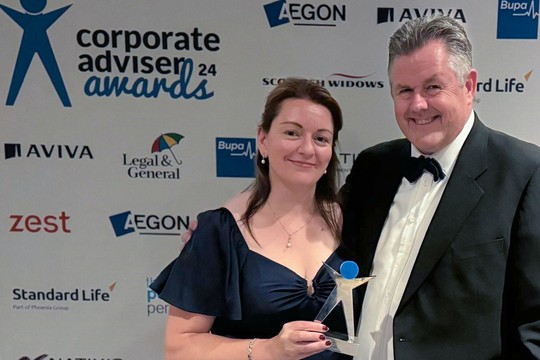 A man and a woman pose together, smiling and holding a trophy, in front of a backdrop with multiple corporate logos, suggesting a formal awards event.