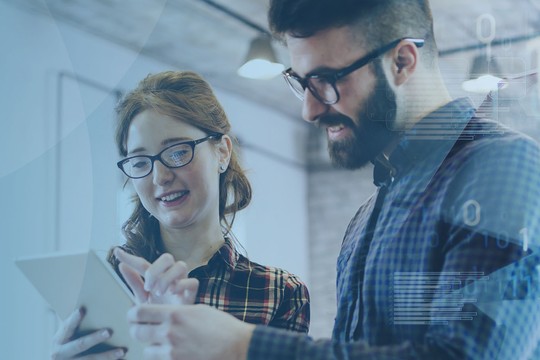 A man and woman are engaged in discussion while reviewing information on a tablet. They are in a modern office space, characterized by large windows and a collaborative atmosphere.