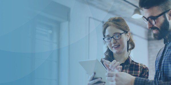 A man and woman are engaged in discussion while reviewing information on a tablet. They are in a modern office space, characterized by large windows and a collaborative atmosphere.