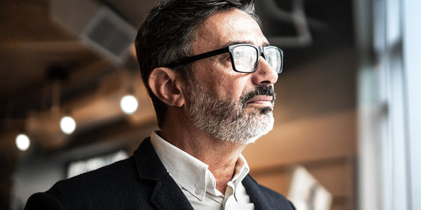 A middle-aged man with glasses and a beard gazes thoughtfully while standing indoors, surrounded by a softly lit modern space featuring blurred backgrounds and hanging light fixtures.