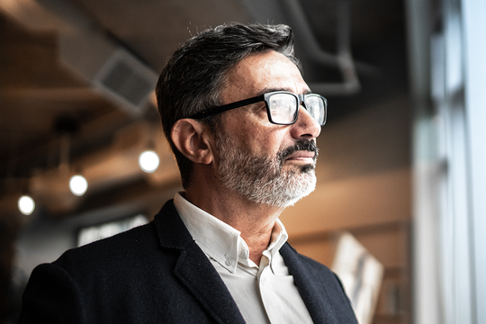 A middle-aged man with glasses and a beard gazes thoughtfully while standing indoors, surrounded by a softly lit modern space featuring blurred backgrounds and hanging light fixtures.
