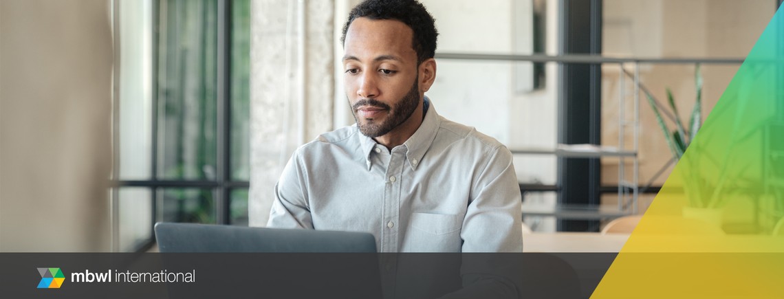 A man sits at a desk, focused on his laptop. He is in a modern workspace with large windows, allowing natural light and greenery to be visible in the background.
