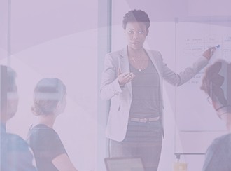 A business professional in a gray blazer gestures while presenting ideas to an audience seated at a table, with a whiteboard in the background in a bright, modern meeting room.